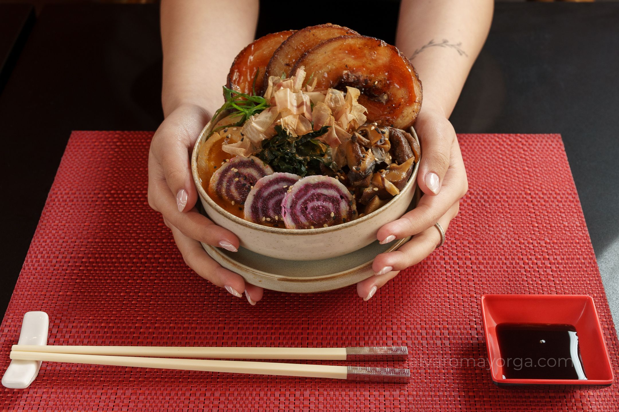 Ramen with pork and vegetables presented in hands over a red table.